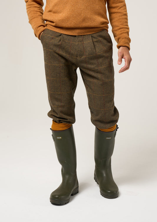 Man wears Combrook Men’s Tweed Water Repellent Shooting Breeks in Teak, tan sweater, green boots against plain backdrop.