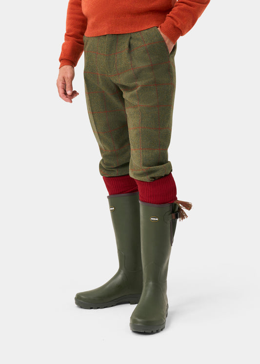 Person in Combrook Men’s Tweed Shooting Breeks (Reed), red socks, olive boots, orange sweater, white background.