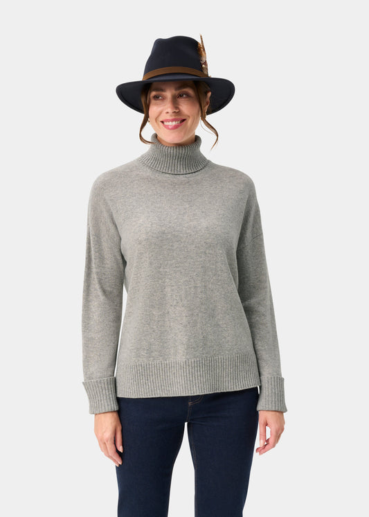 Woman in the Alexa Wool & Cashmere Roll Neck Jumper in Husky, smiling in a dark feathered hat against a white background.