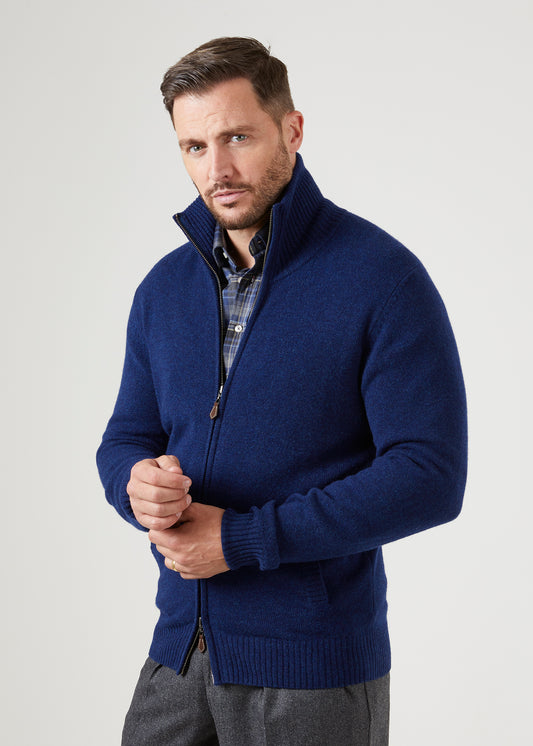 Man in a Ballater Lambswool Front Zip Mock Neck Jumper in Indigo, hands clasped, looking forward neutrally.