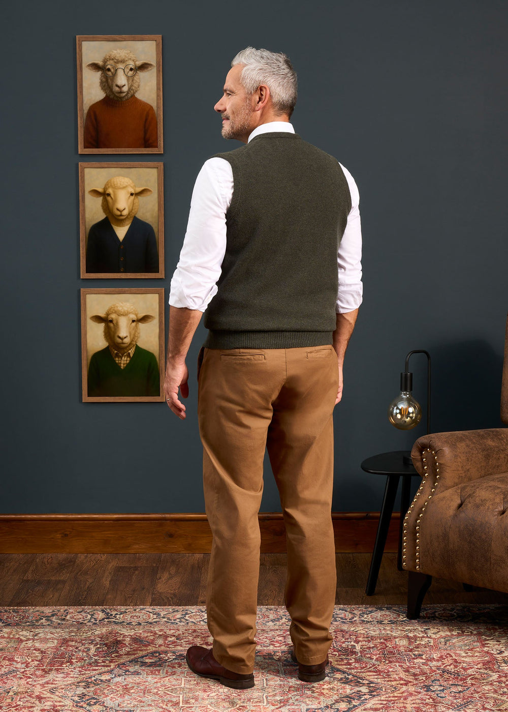 A man wears a Norfolk Lambswool V-Neck Slipover In Seaweed over a white shirt and tan trousers, seen from behind.