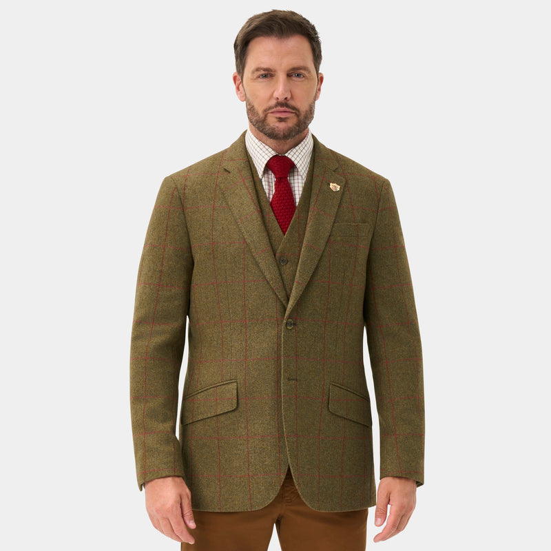 A man modeling a tailored suit featuring an olive green tweed blazer, matching brown waistcoat and trousers, a white dress shirt, a red patterned tie, and dark brown leather shoes, set against a plain light gray background.