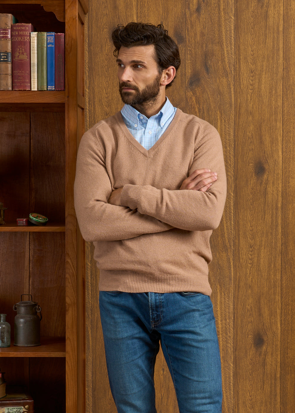 Man in a Camel Hampshire Lambswool Vee Neck Jumper and jeans stands by a wood bookshelf and paneled wall.