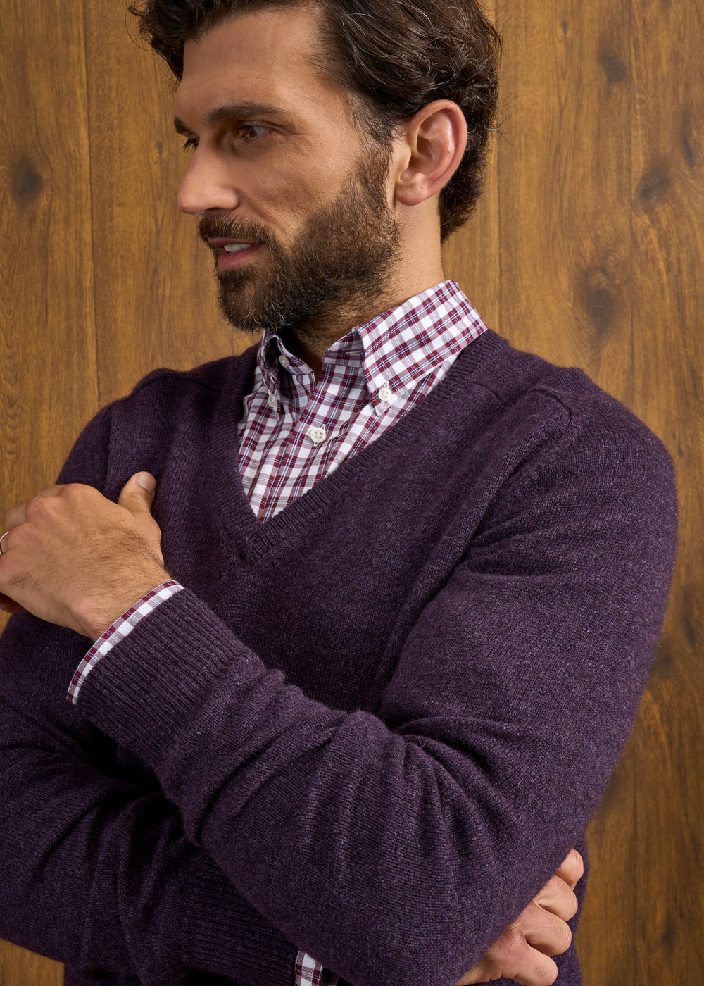 Man with brown hair and beard in a Hampshire Lambswool Vee Neck Jumper in Elderberry, by a wooden wall.