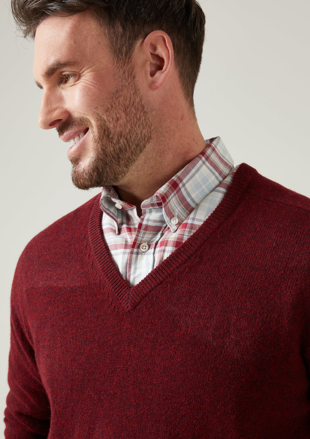 Man smiles in Hampshire Lambswool Vee Neck Jumper, Red Velvet, Classic Fit, over plaid shirt, facing left.