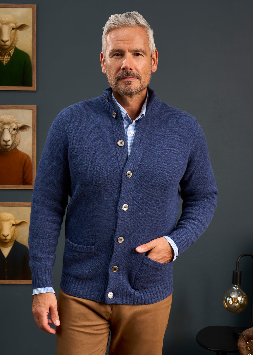 A man wears the Landford Lambswool Buttoned Mock Neck Jumper in Rhapsody, indoors with sheep art and a lamp nearby.