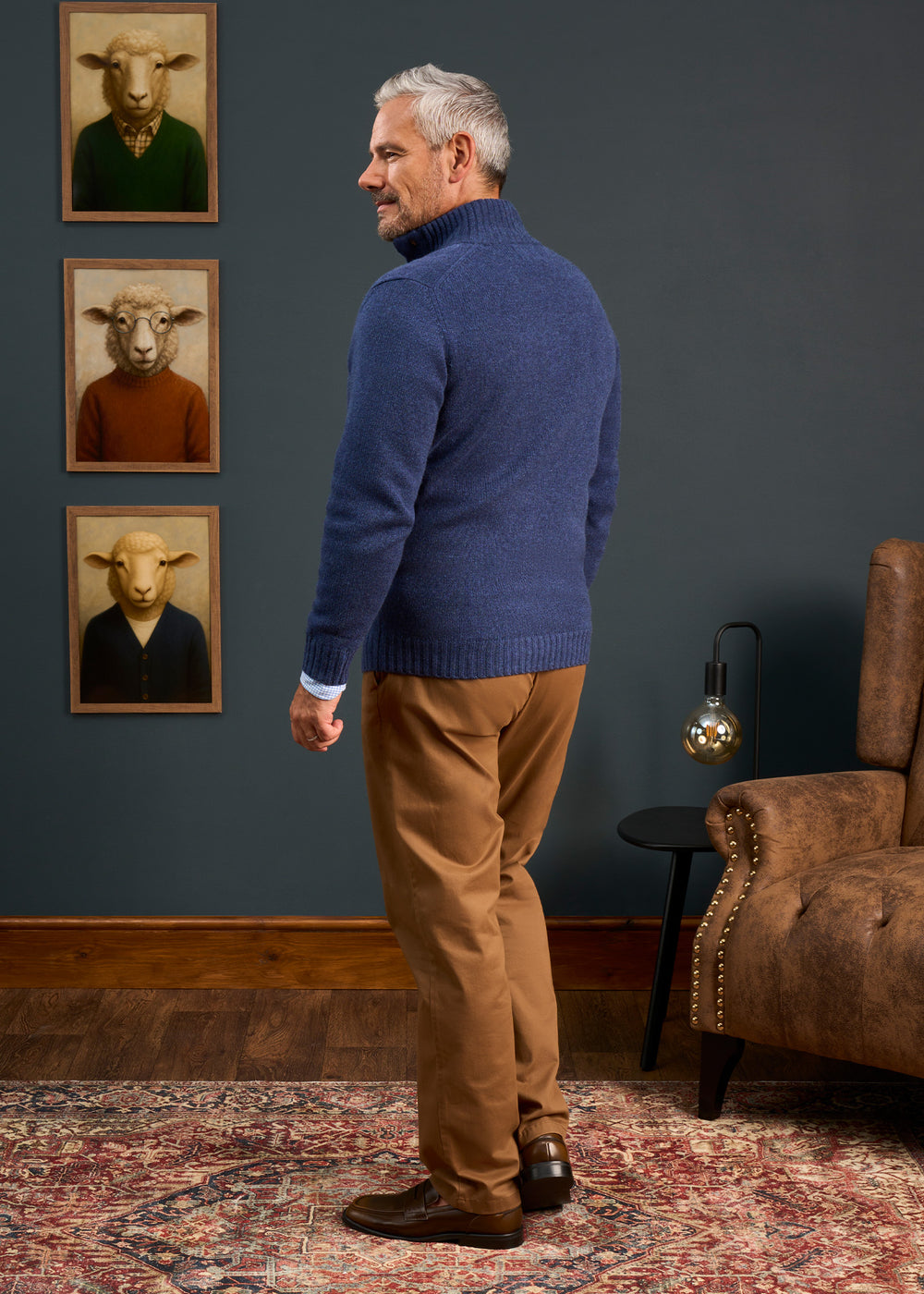 Man wears Landford Lambswool Buttoned Mock Neck Jumper in Rhapsody, tan pants; sheep art, brown chair behind him.