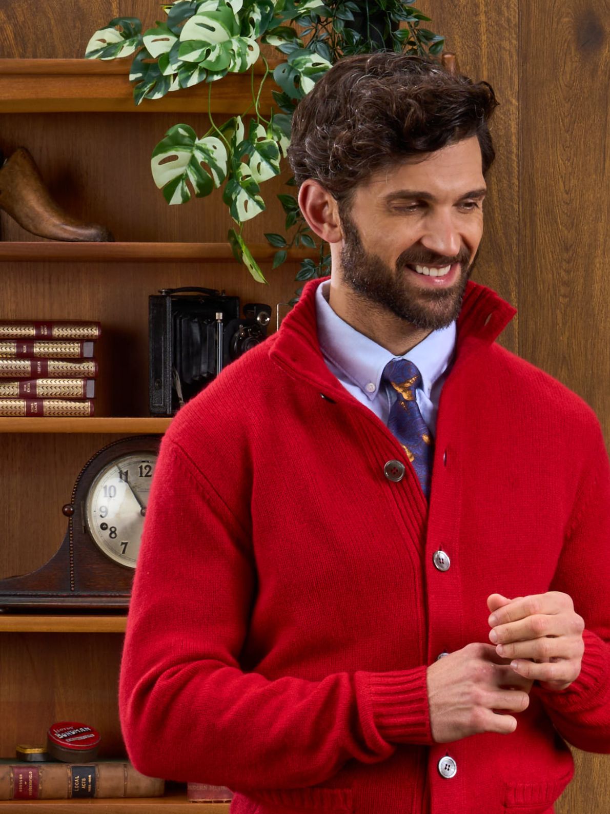 Smiling man in a Landford Lambswool Jumper stands before shelves with books, a clock, and a plant.