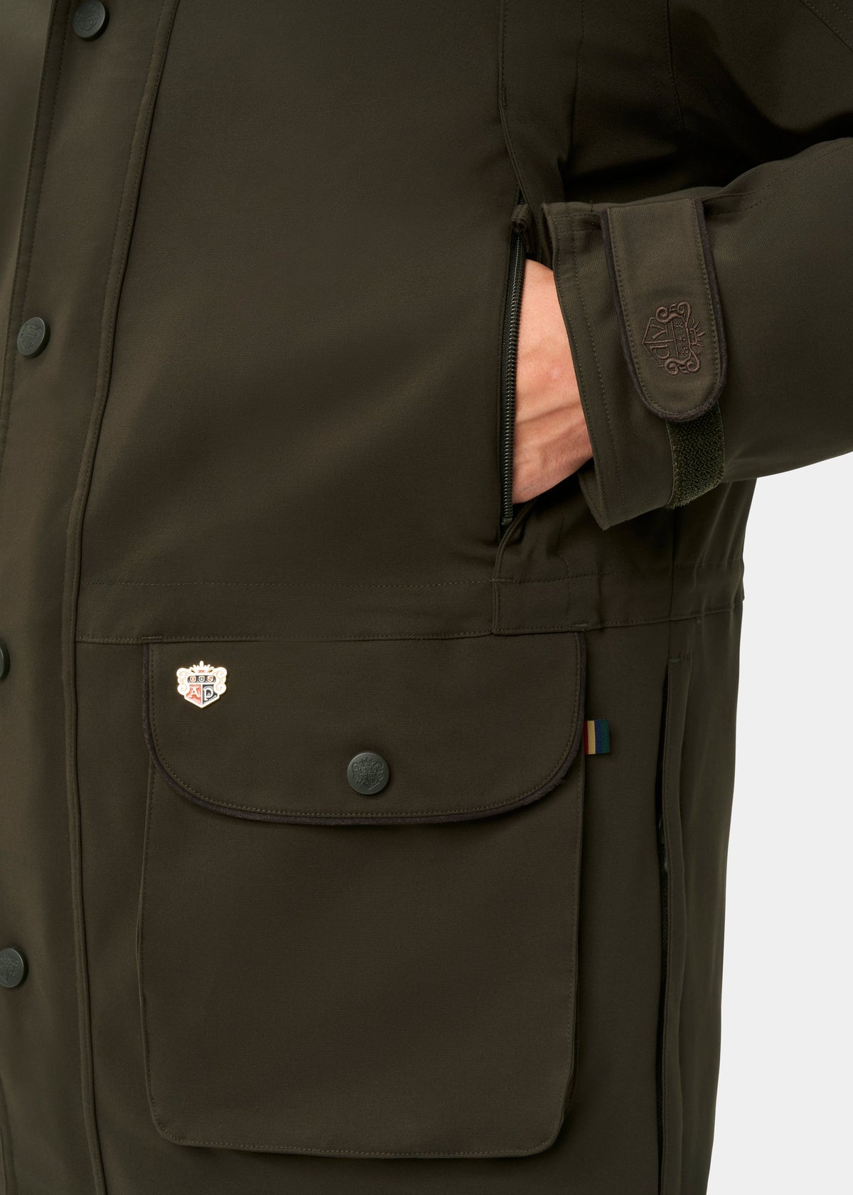 A hand in the pocket of a Dunswell Men’s Waterproof Field Coat in Olive with buttons, patches, and a velcro wrist strap.