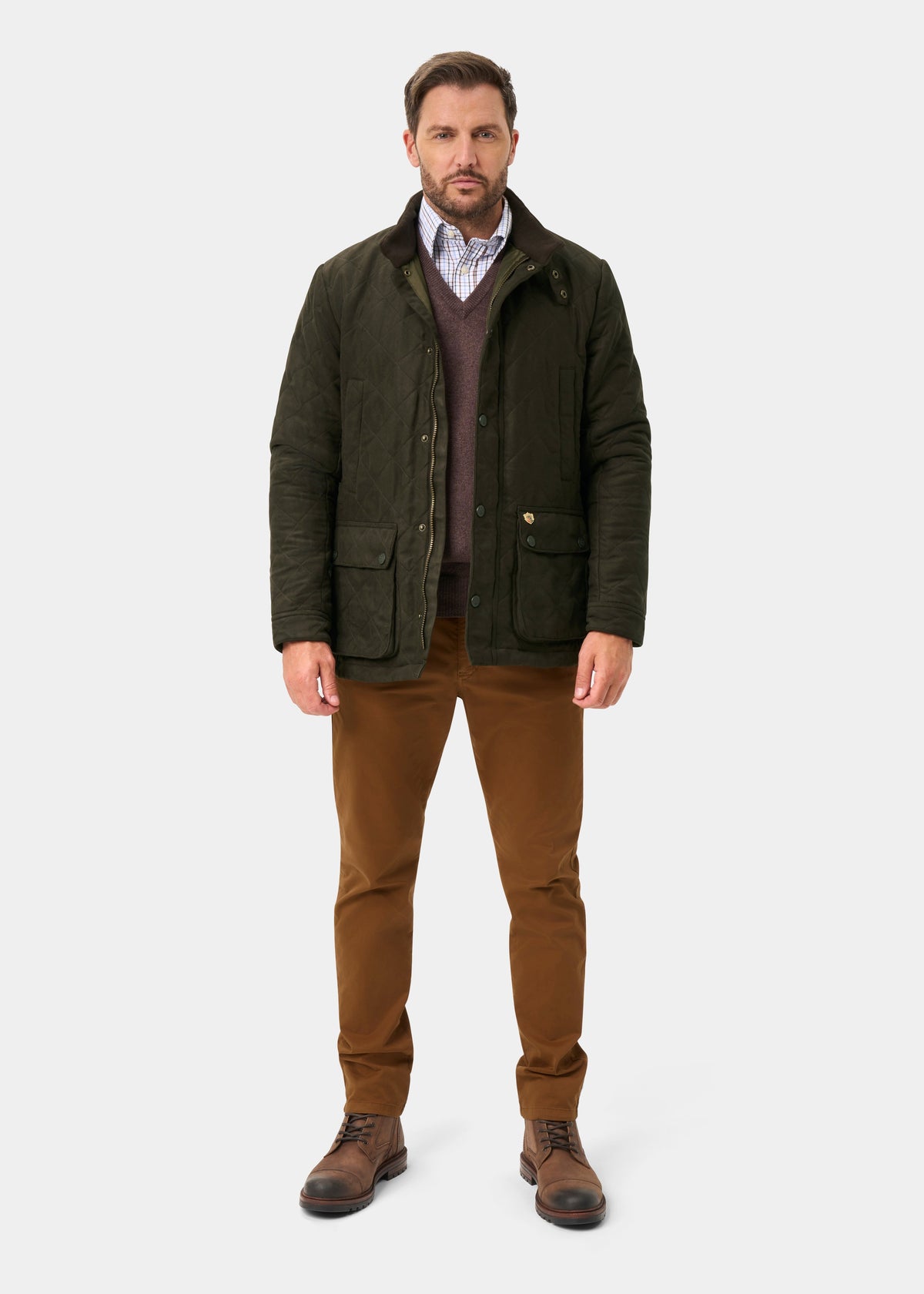 Man wearing Felwell Men’s Quilted Jacket in Olive, brown pants, boots, and v-neck sweater over collared shirt.