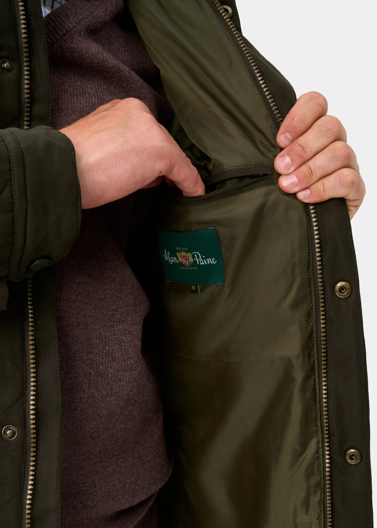 A hand reaches into the inner pocket of the Felwell Men’s Quilted Jacket in Olive, revealing its brand label.