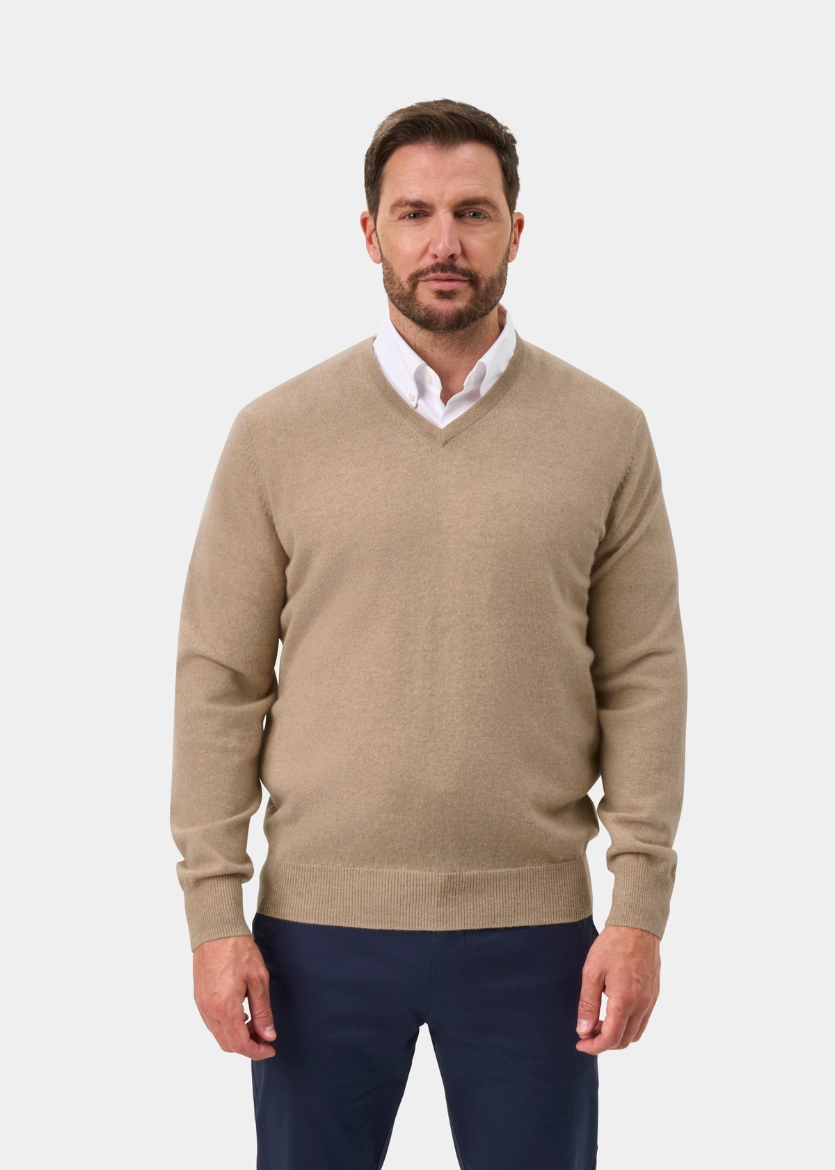 A man wears the Haddington Pure Cashmere V-Neck Jumper in Linen over a white shirt and navy pants, on a white background.