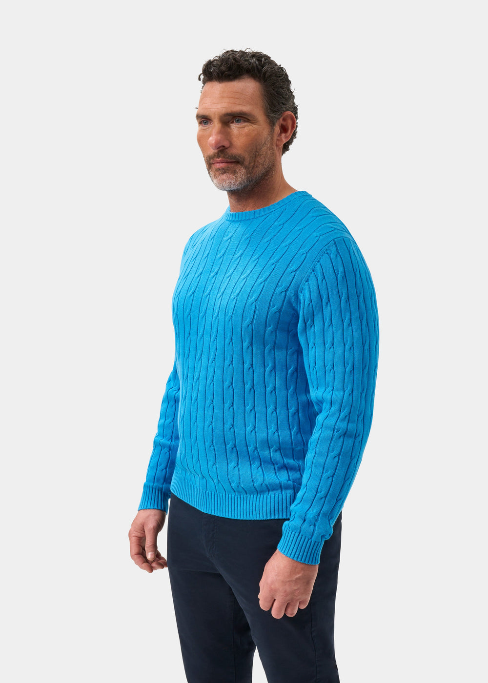 Man wears Honley Cotton Cashmere Cable Knit Crew Neck Jumper in Blue Lagoon, with dark pants, against white background.