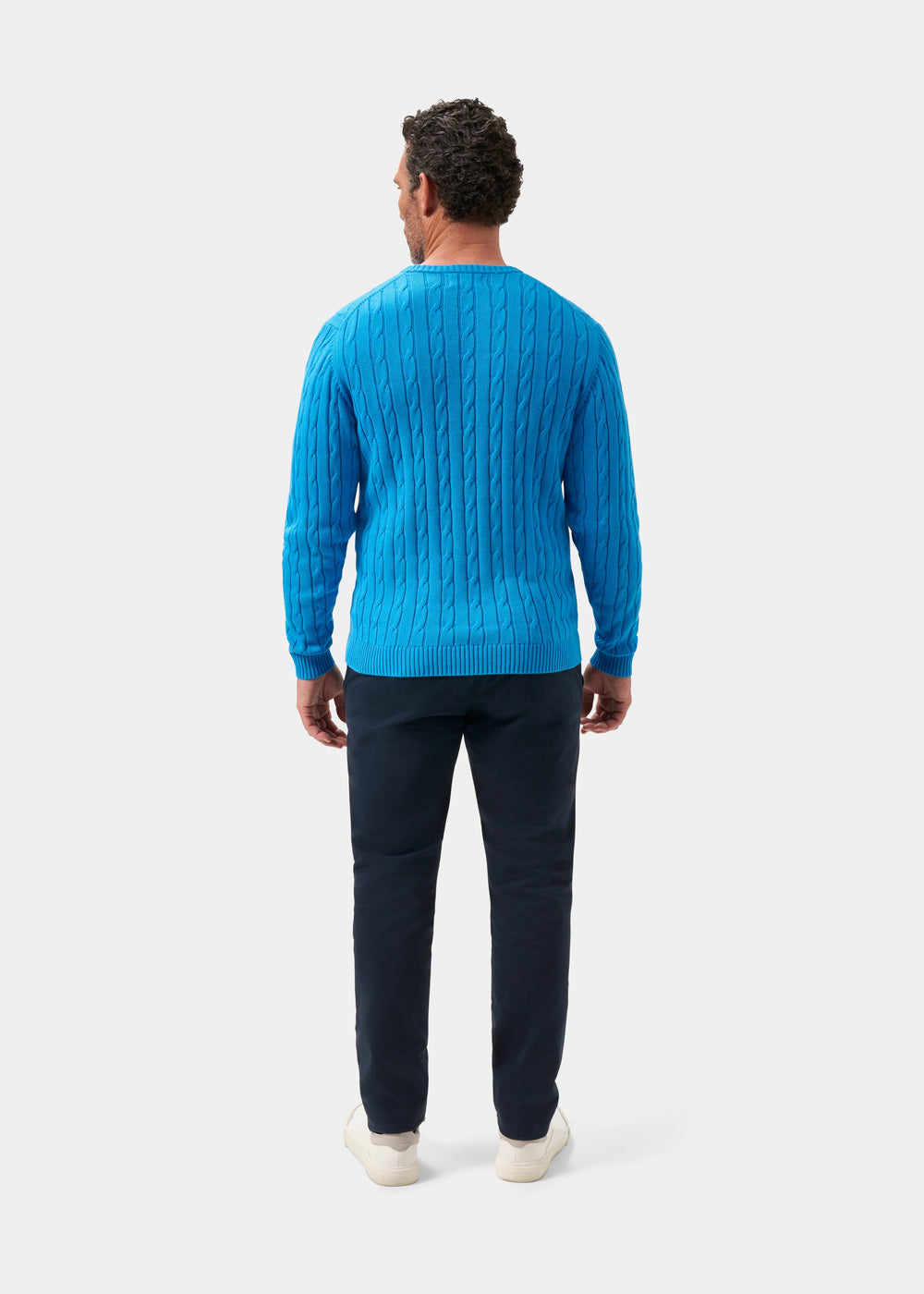 Man facing away in Honley Cotton Cashmere Cable Knit Crew Neck Jumper Blue Lagoon, dark pants, white shoes, plain backdrop.