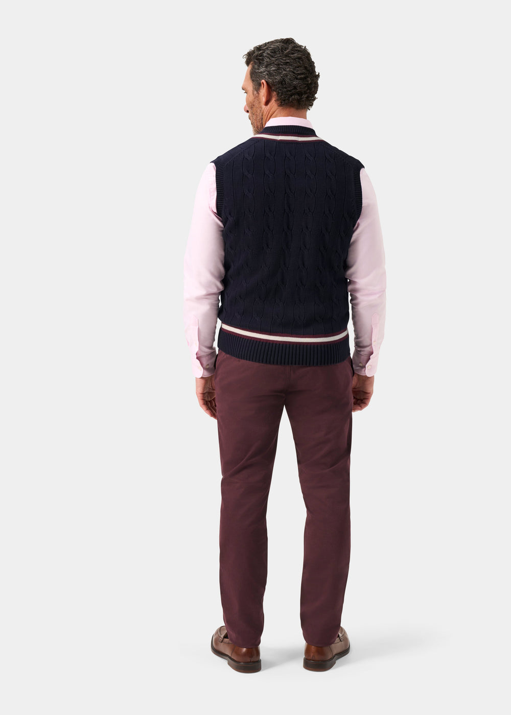 Man facing away in a pink shirt and Chadbury Navy & Blackcurrant Cricket Slipover, maroon pants, brown shoes.