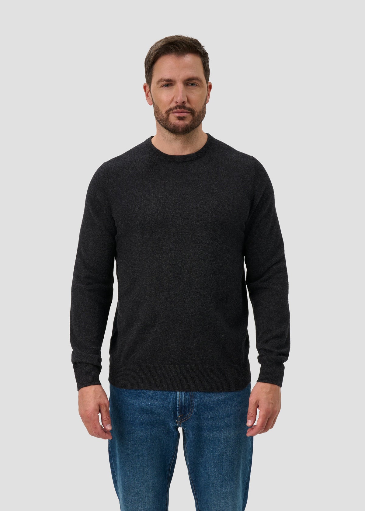 Man with short brown hair and beard wears Brisbane Geelong Wool Crew Neck Jumper in charcoal and blue jeans.