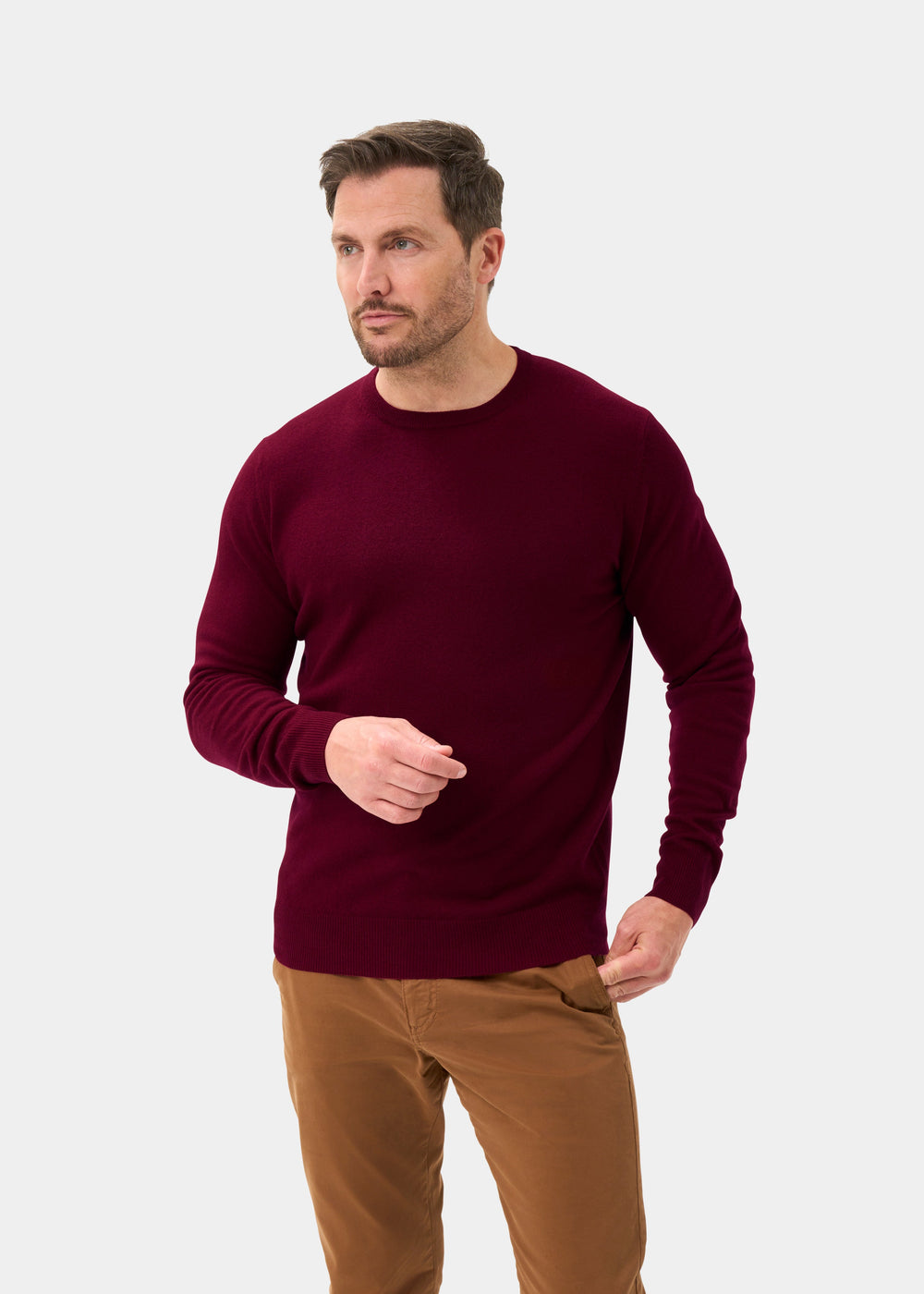 Man in Brisbane Geelong Wool Crew Neck Jumper in Claret and tan pants stands against a light gray background.