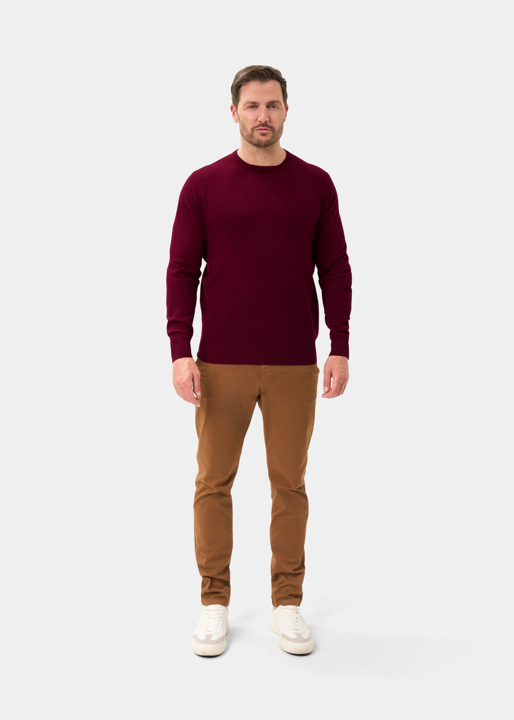 Man in a claret Brisbane Geelong wool crew neck jumper, tan pants, and white sneakers on a light gray background.