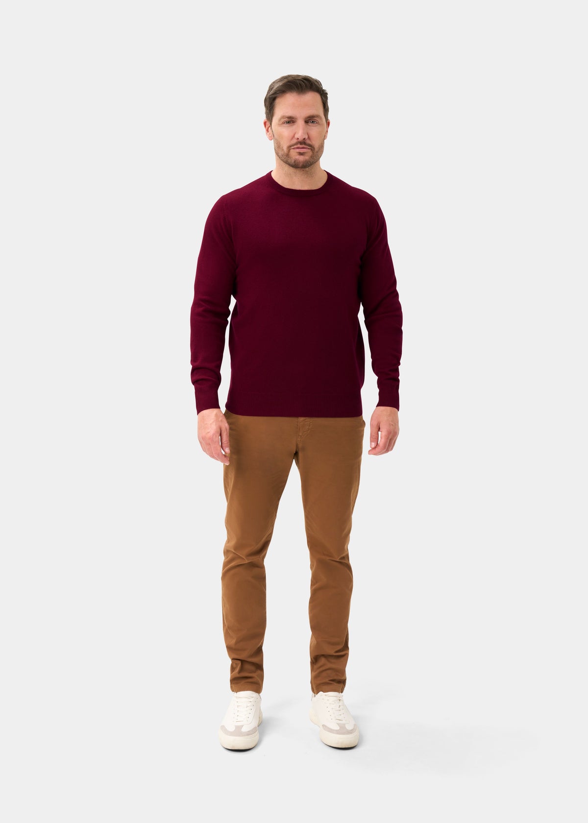 Man in a claret Brisbane Geelong wool crew neck jumper, tan pants, and white sneakers on a light gray background.