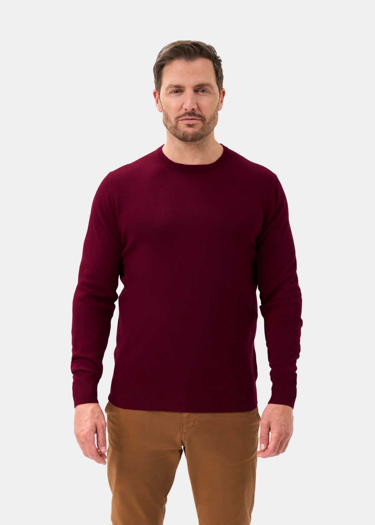 Man in a Brisbane Geelong Wool Crew Neck Jumper in Claret and brown pants stands against a light gray background.