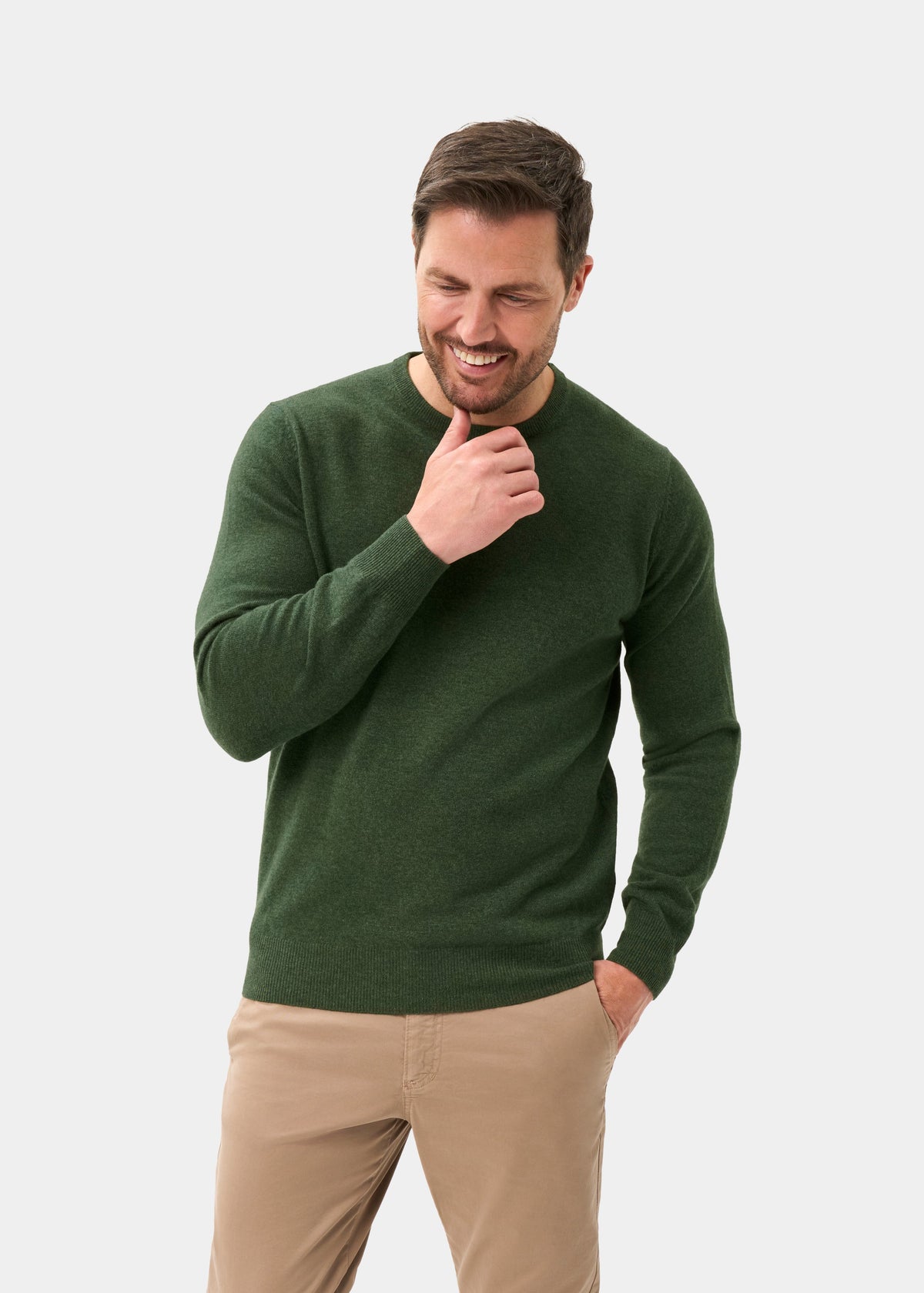 Man smiles in a rosemary Brisbane Geelong Wool Crew Neck Jumper and beige pants, hand on chin, other in pocket.