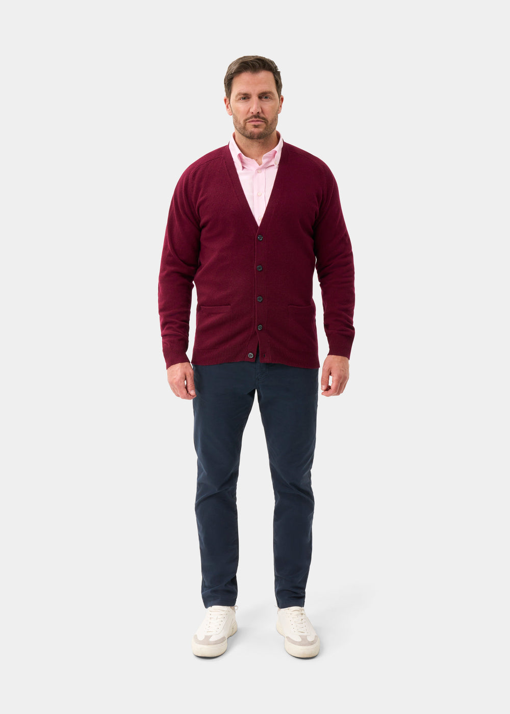 Man in a Cornwall Lambswool V-Neck Cardigan in Bordeaux, pink shirt, and navy pants stands against a gray background.