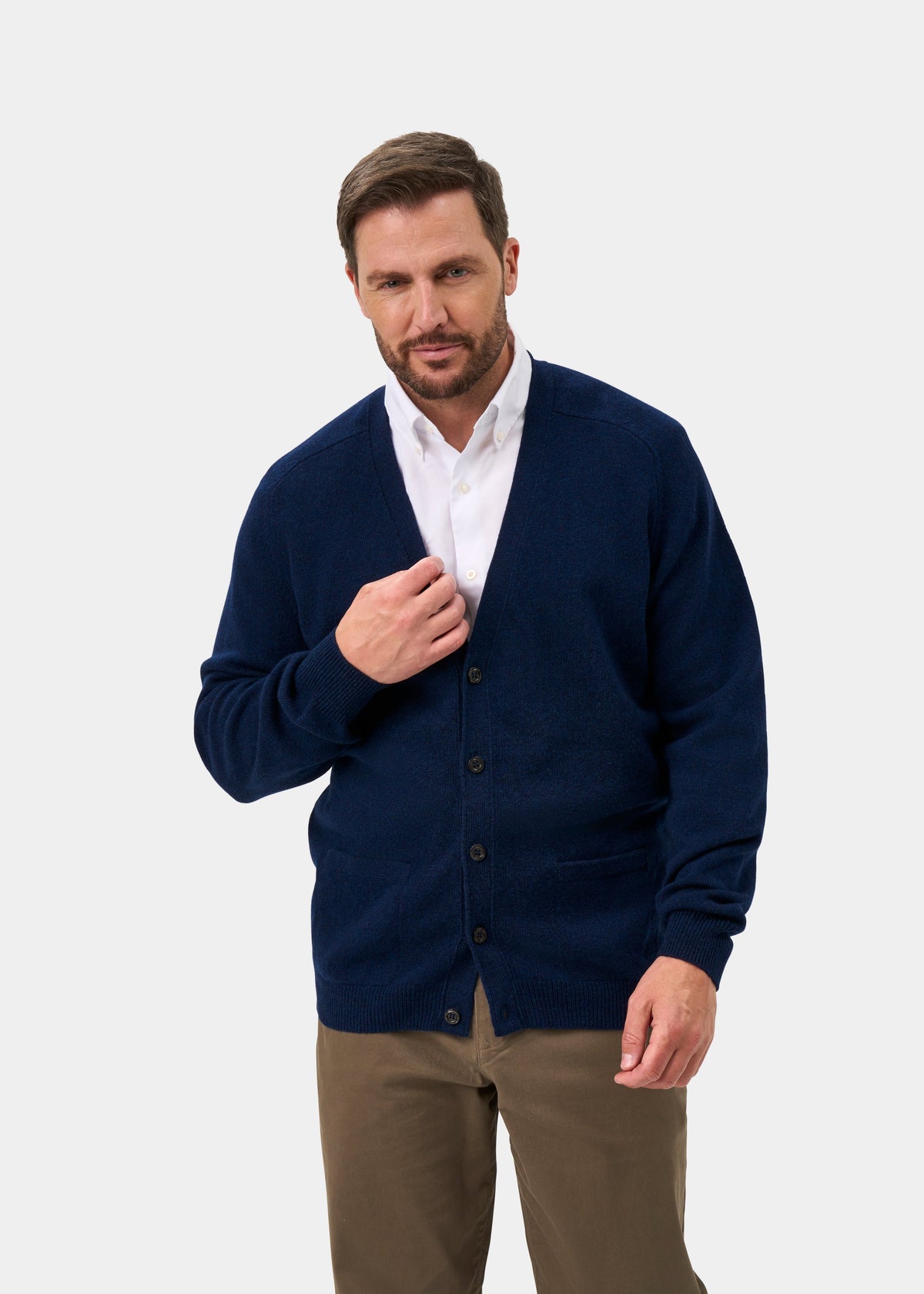 Man in Cornwall Lambswool V-Neck Cardigan in Indigo, white shirt, brown pants, on plain light background.