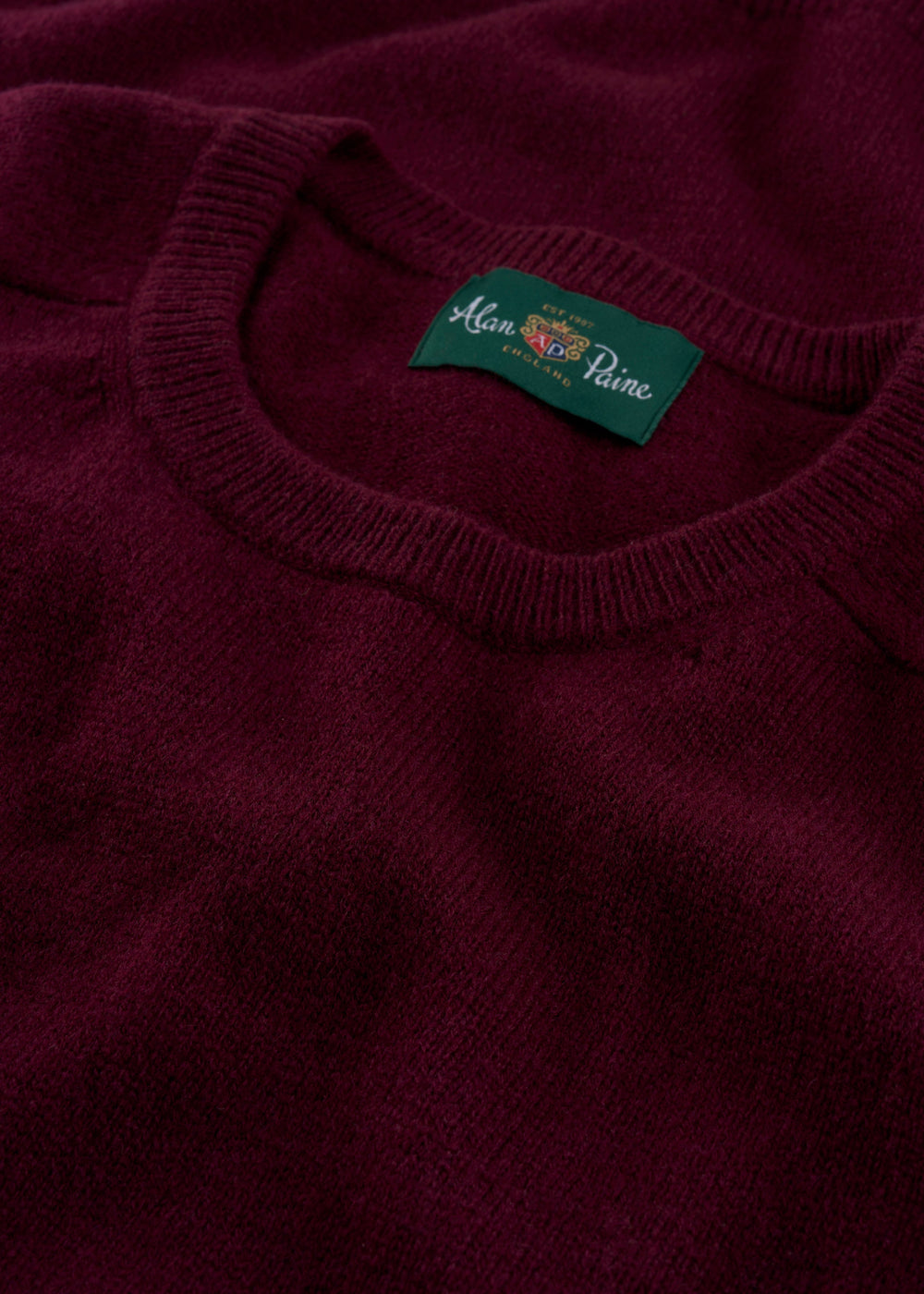 Close-up of the Lenzie Lambswool Crew Neck Jumper in Bordeaux with a green Alan Paine England label on the collar.