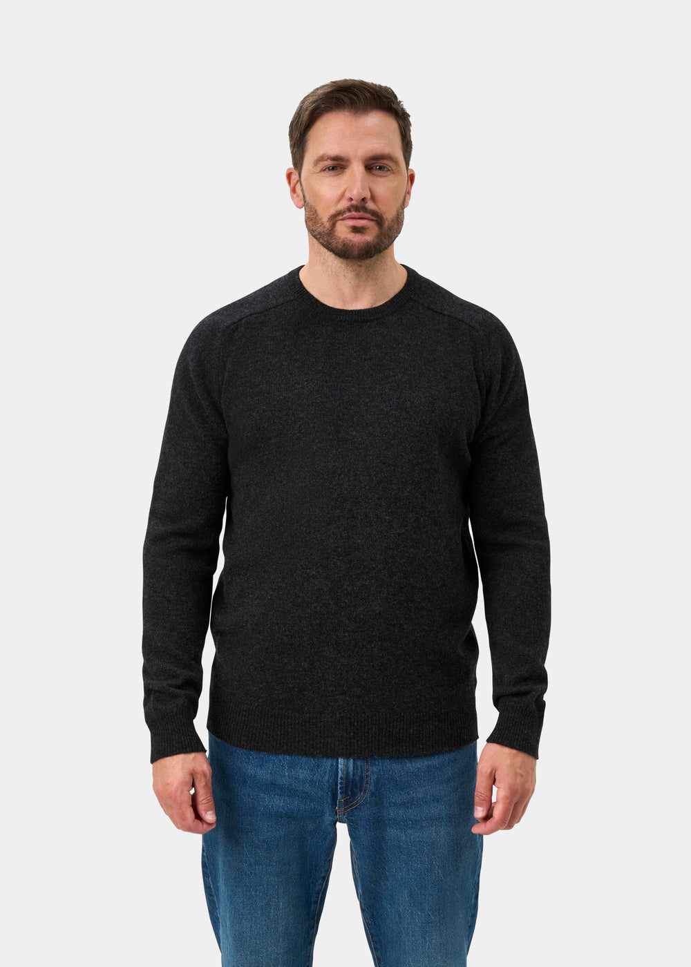 Man wears Lenzie Lambswool Crew Neck Jumper in Charcoal, regular fit, with blue jeans against a white background.