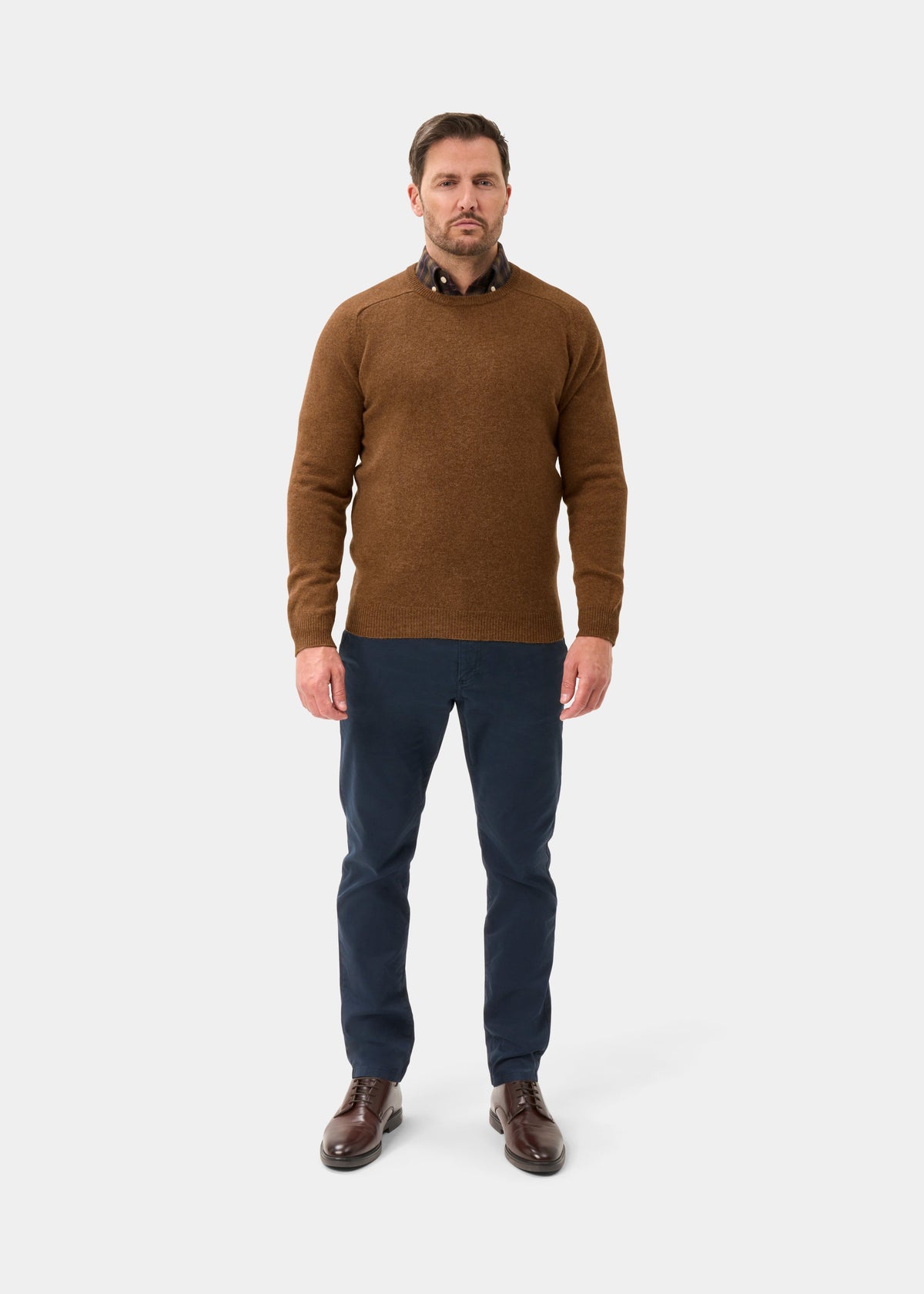 Man in Dorset Lambswool Crew Neck Jumper in Driftwood, blue pants, and brown shoes against a plain background.
