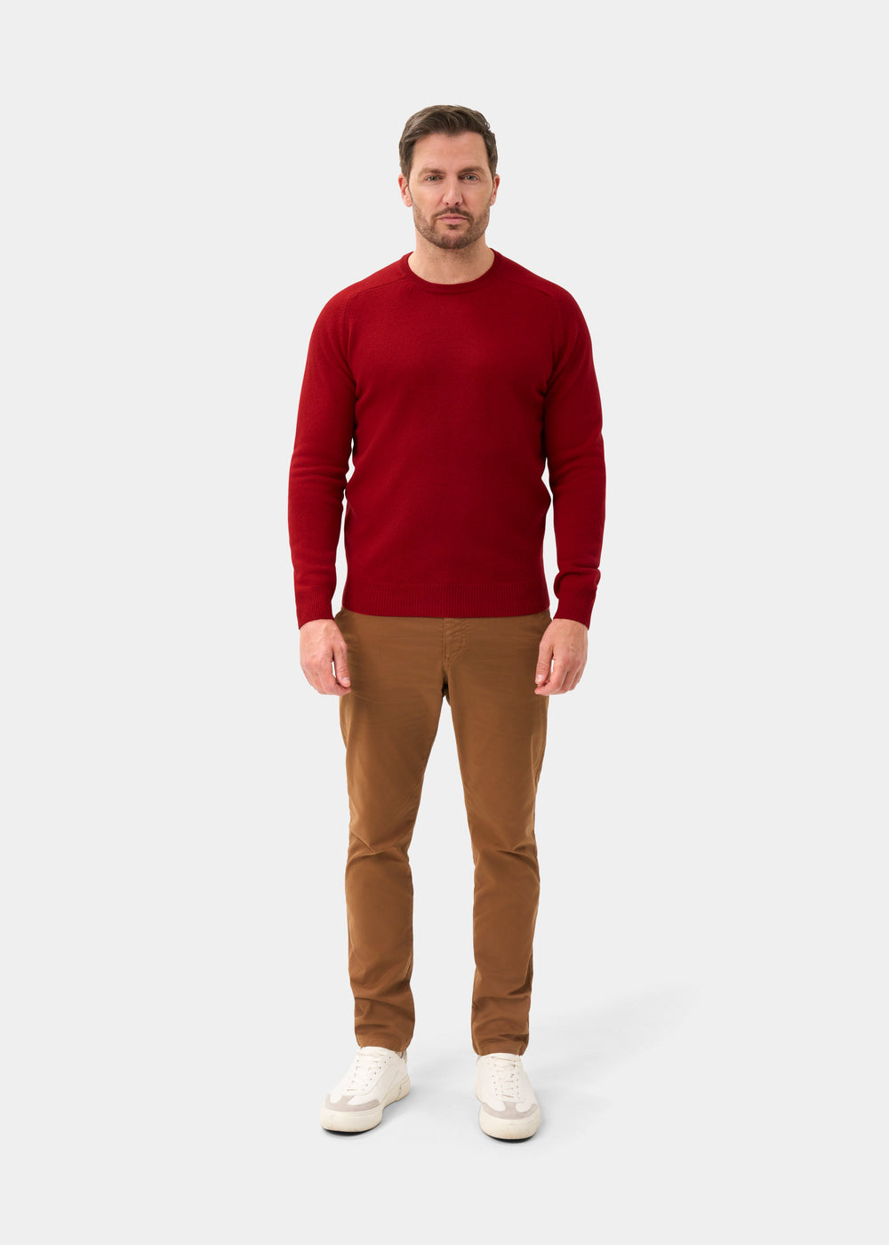 Man in a Lenzie Lambswool Crew Neck Jumper in Dubonnet, tan pants, and white sneakers on a plain light gray background.