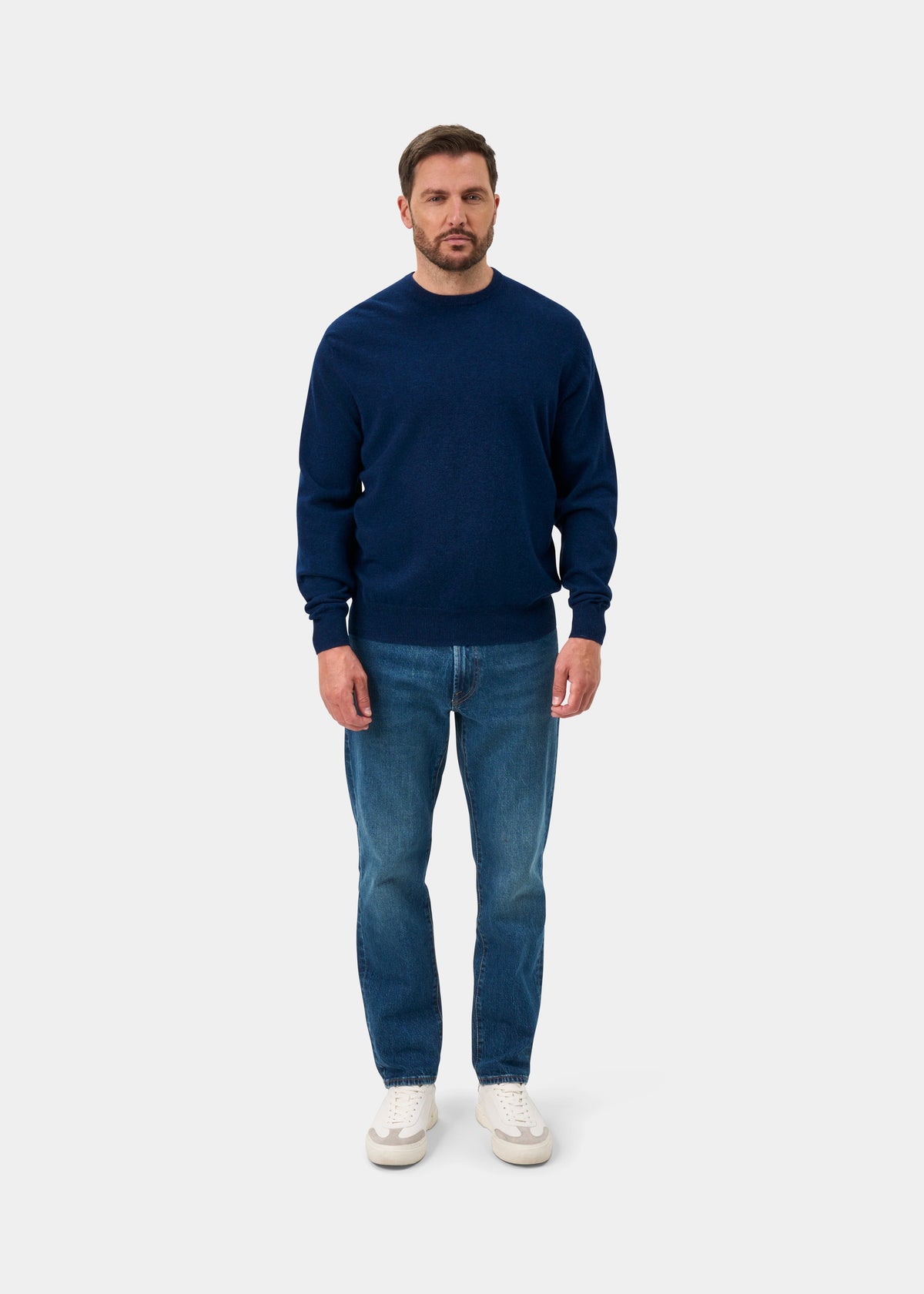 Man in Dorset Lambswool Crew Neck Jumper in Indigo, blue jeans, white sneakers stands facing forward on white background.
