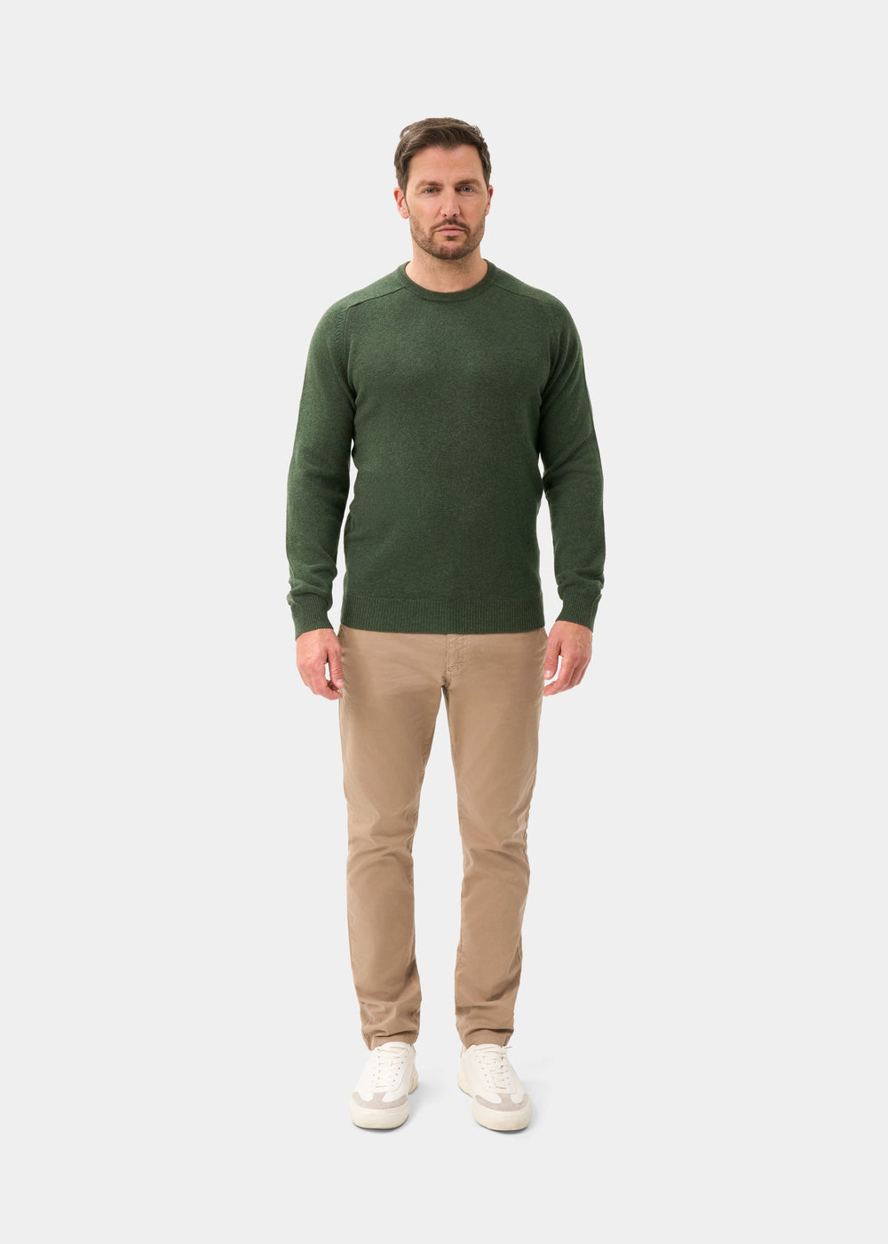 Man wears Dorset Lambswool Crew Neck Jumper in Rosemary, beige pants, and white sneakers on a plain background.