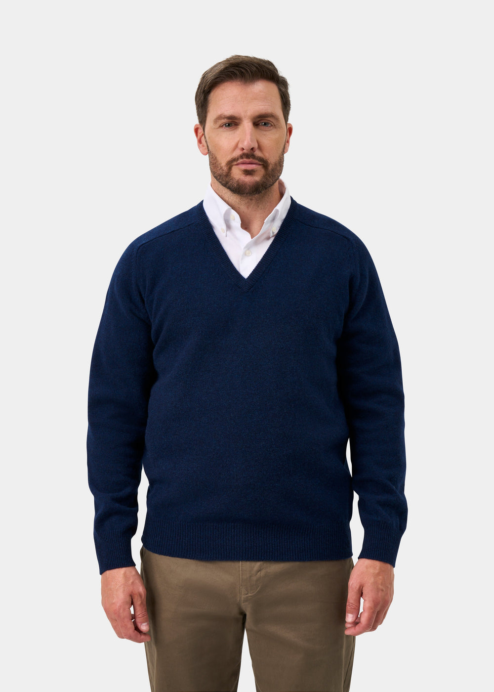 Man in Hampshire Lambswool V-Neck Jumper in Indigo over a white shirt and khaki pants stands against a plain backdrop.