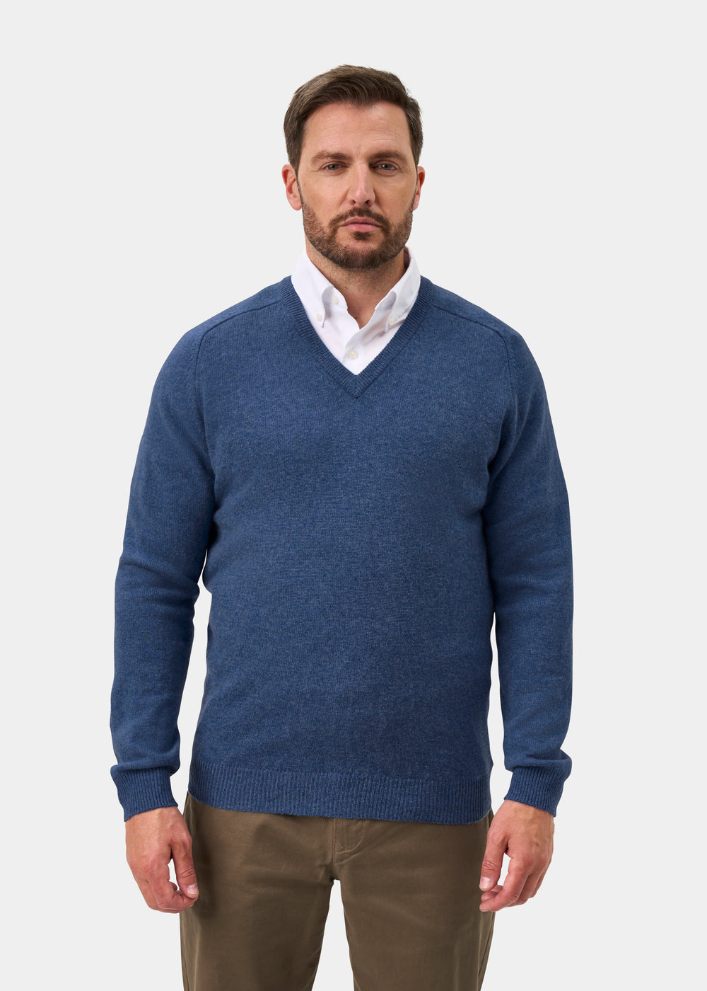 A bearded man wears the Kilsyth Lambswool Vee Neck Jumper in Jeans over a white shirt and brown pants against white.