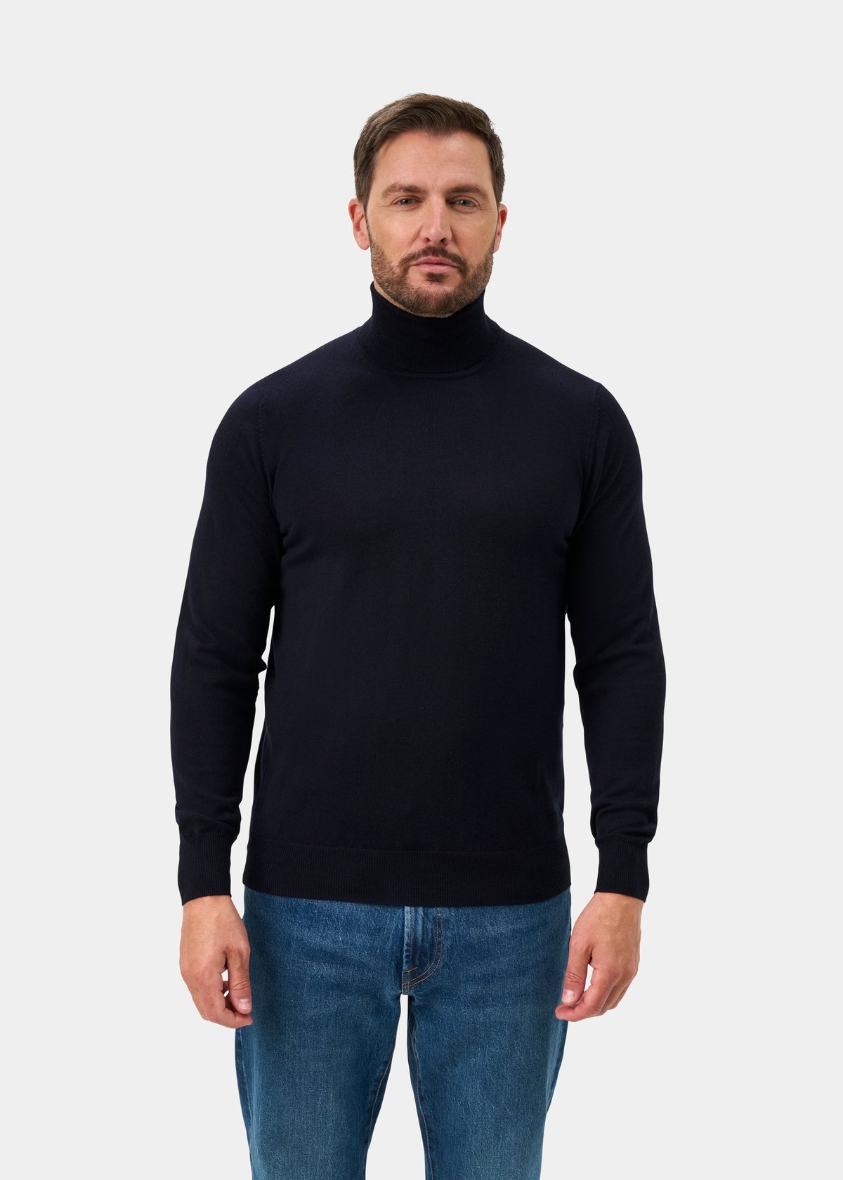 Man wears Linton Merino Wool Roll Neck Jumper in Dark Navy, paired with blue jeans against a plain background.