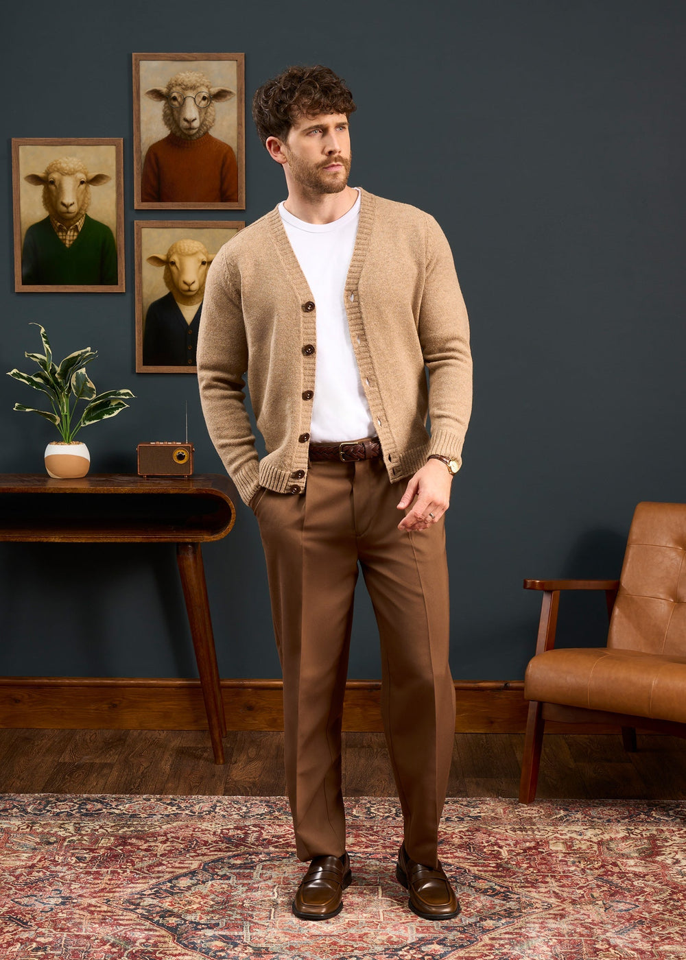 Man in Kinloch Supersoft Shetland Wool V-Neck Cardigan, Pale Oak, stands with brown pants in a stylish room.