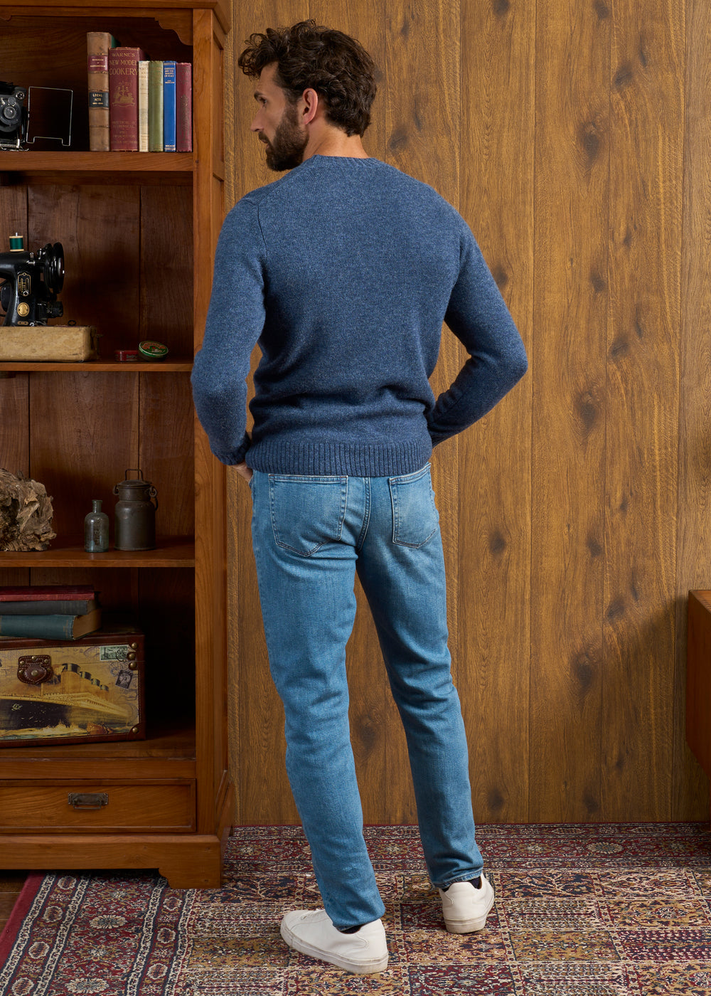 Man in Kinnadie Shetland Wool Crew Neck Jumper (Denim, Regular Fit) and jeans stands by bookshelf on patterned rug.