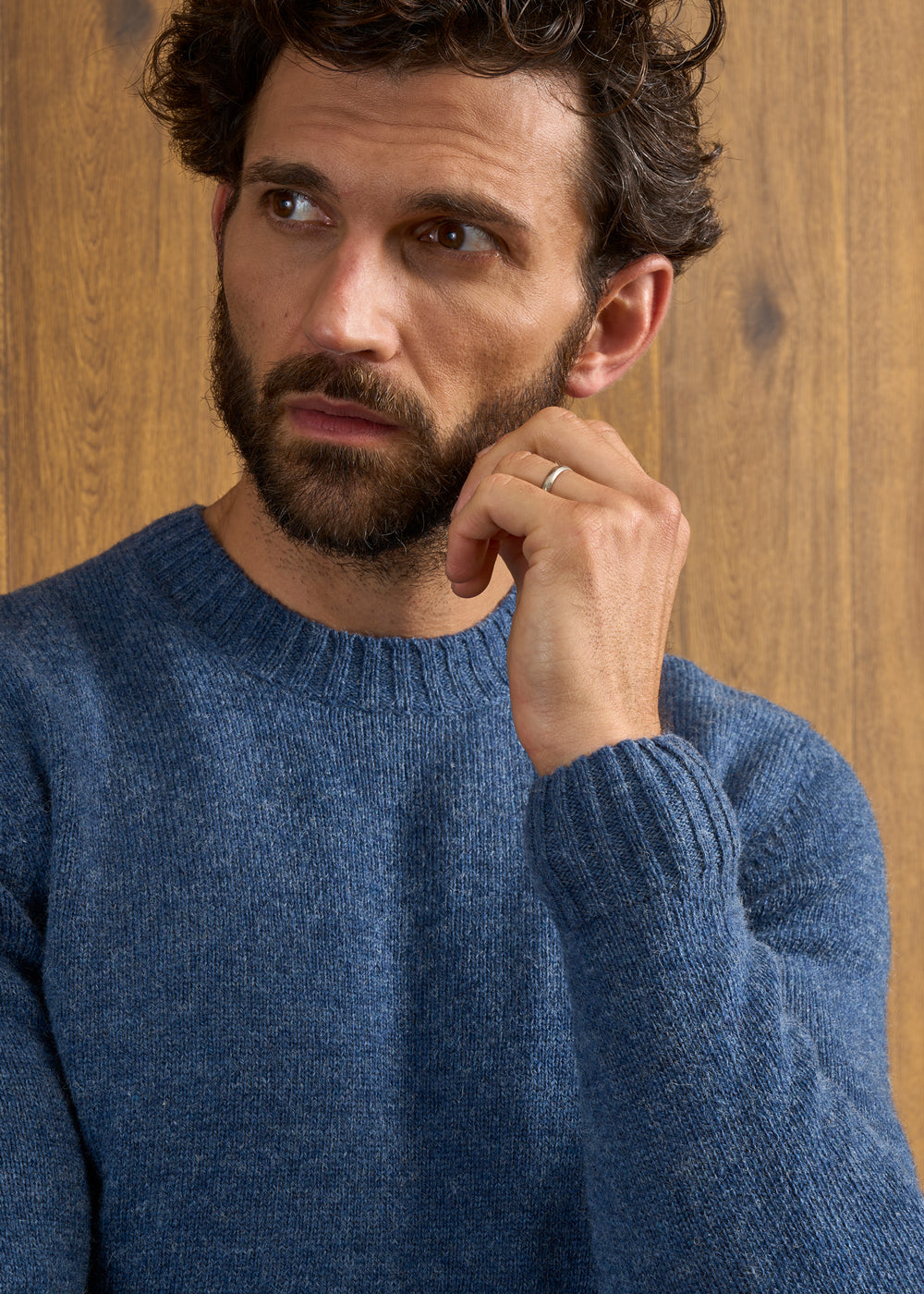 Bearded man in a Kinnadie Supersoft Shetland Wool Crew Neck Jumper in Denim touches his face against wood backdrop.