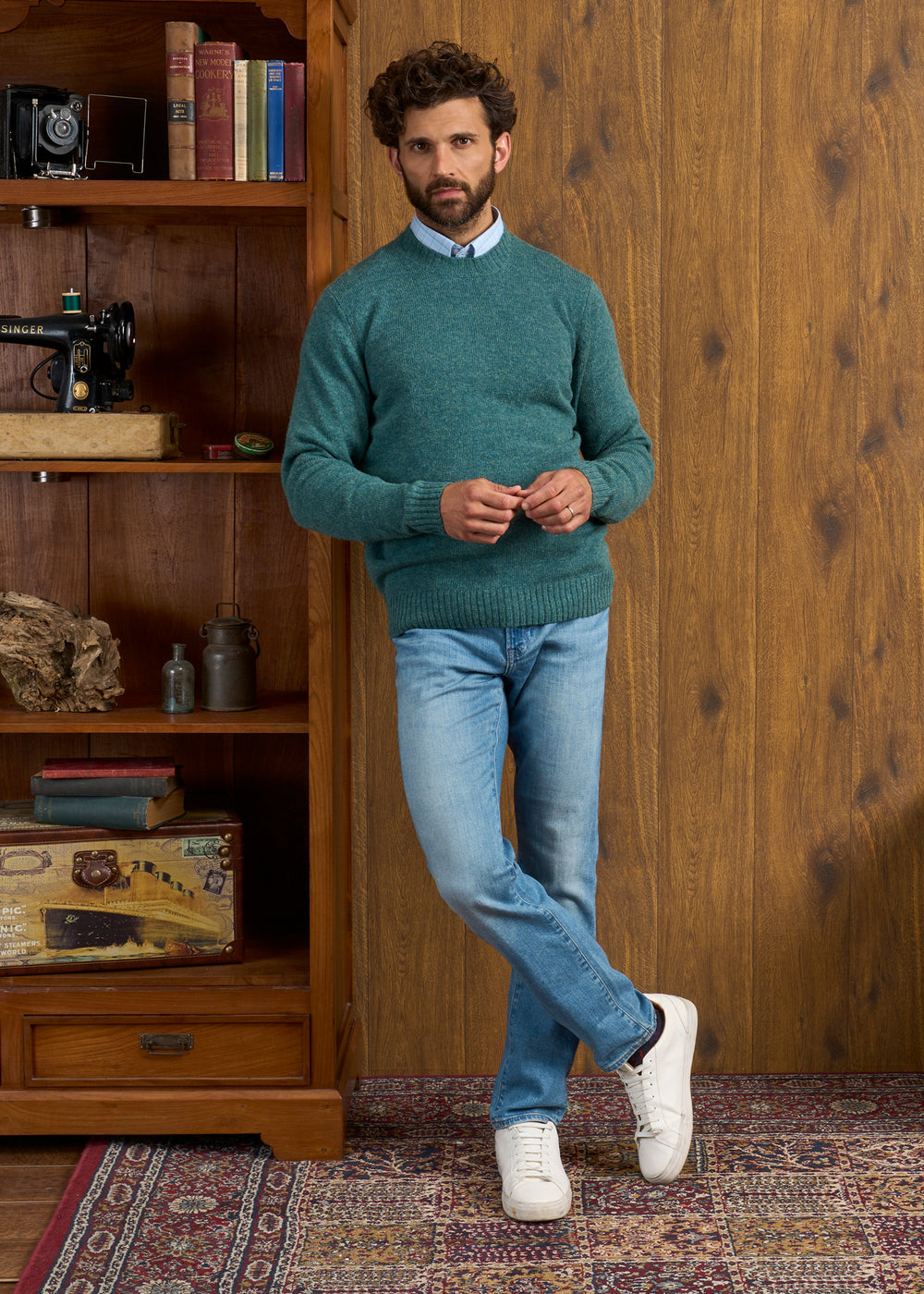 Man in a Kinnadie Supersoft Shetland Wool Jumper leans on a bookcase beside a patterned rug in a cozy room.