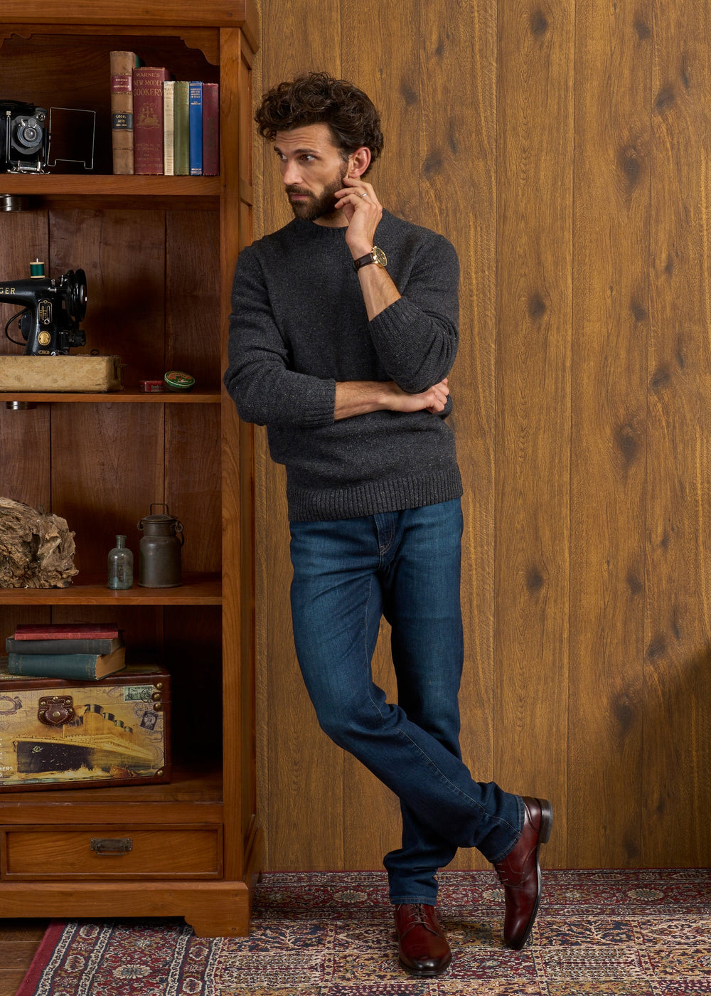 Man in a Kinnadie Shetland Wool Jumper in Oxford Pine and jeans leans on a bookshelf, standing on a patterned rug.