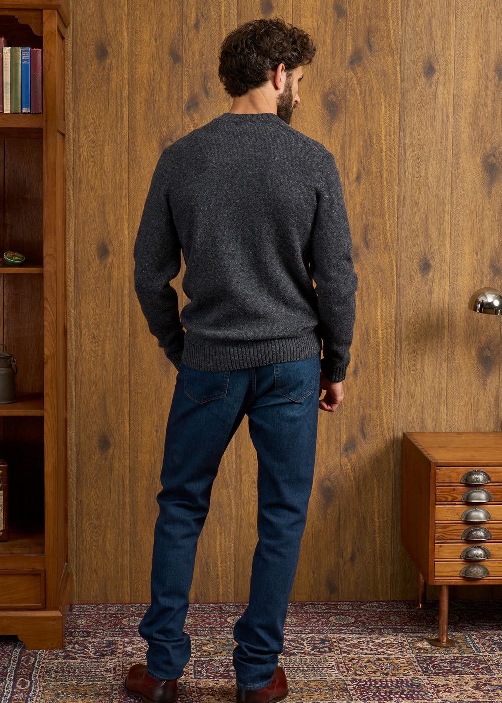 A man wears the Kinnadie Supersoft Shetland Wool Jumper in Oxford Pine, facing a wood-paneled wall with vintage furniture.