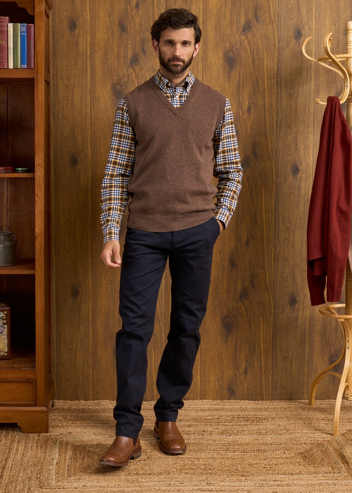 A man wears a Norfolk Lambswool V-Neck Slipover In Tobacco over a plaid shirt, dark trousers, and brown leather boots.