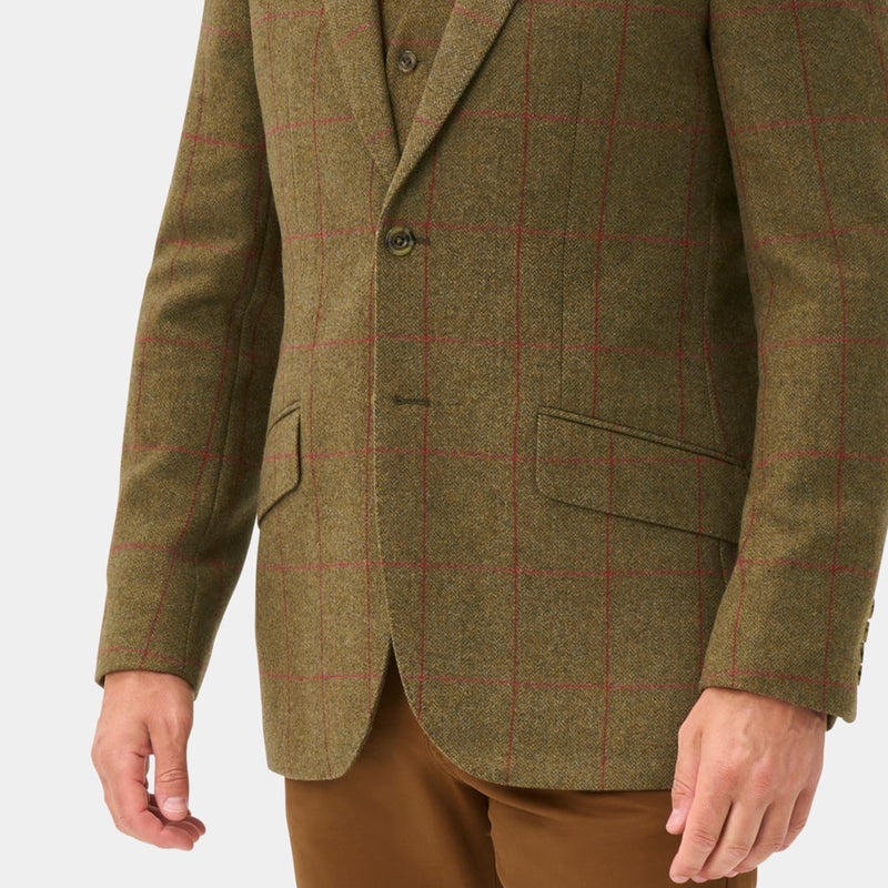 A man modeling a brown tweed sport coat with a subtle plaid pattern, paired with brown trousers, a white shirt, and a red tie, standing against a light gray studio background.