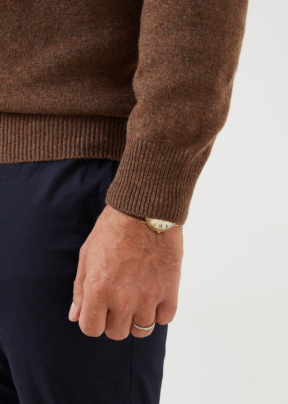 A person in the Dorset Lambswool Crew Neck Jumper in Tobacco, navy pants, gold wristwatch, and a silver ring.