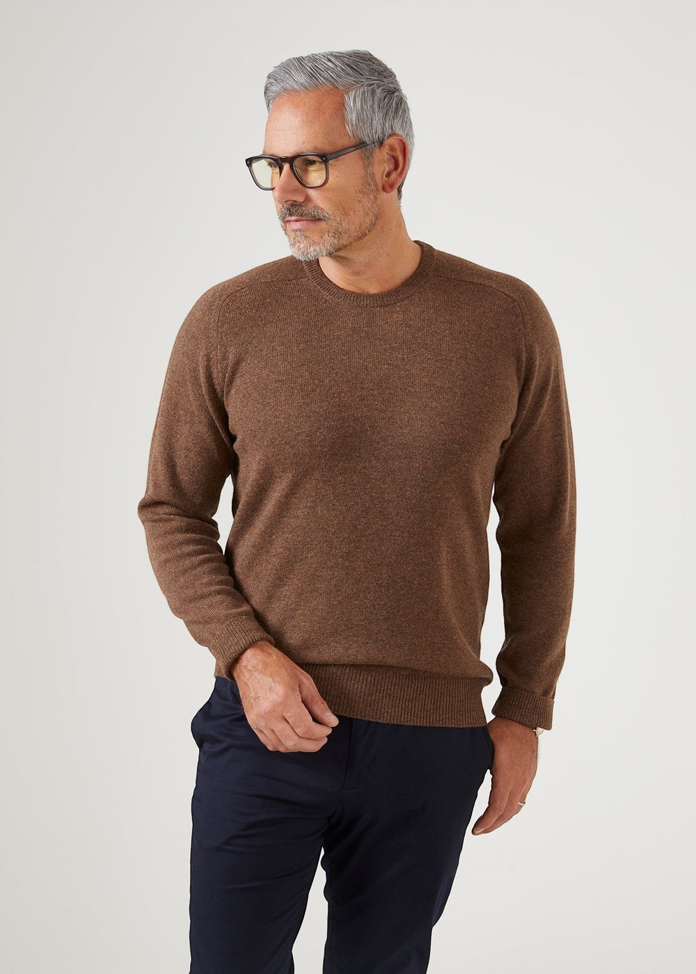 Man with gray hair and glasses wears Dorset Lambswool Crew Neck Jumper in Tobacco and navy pants against white background.