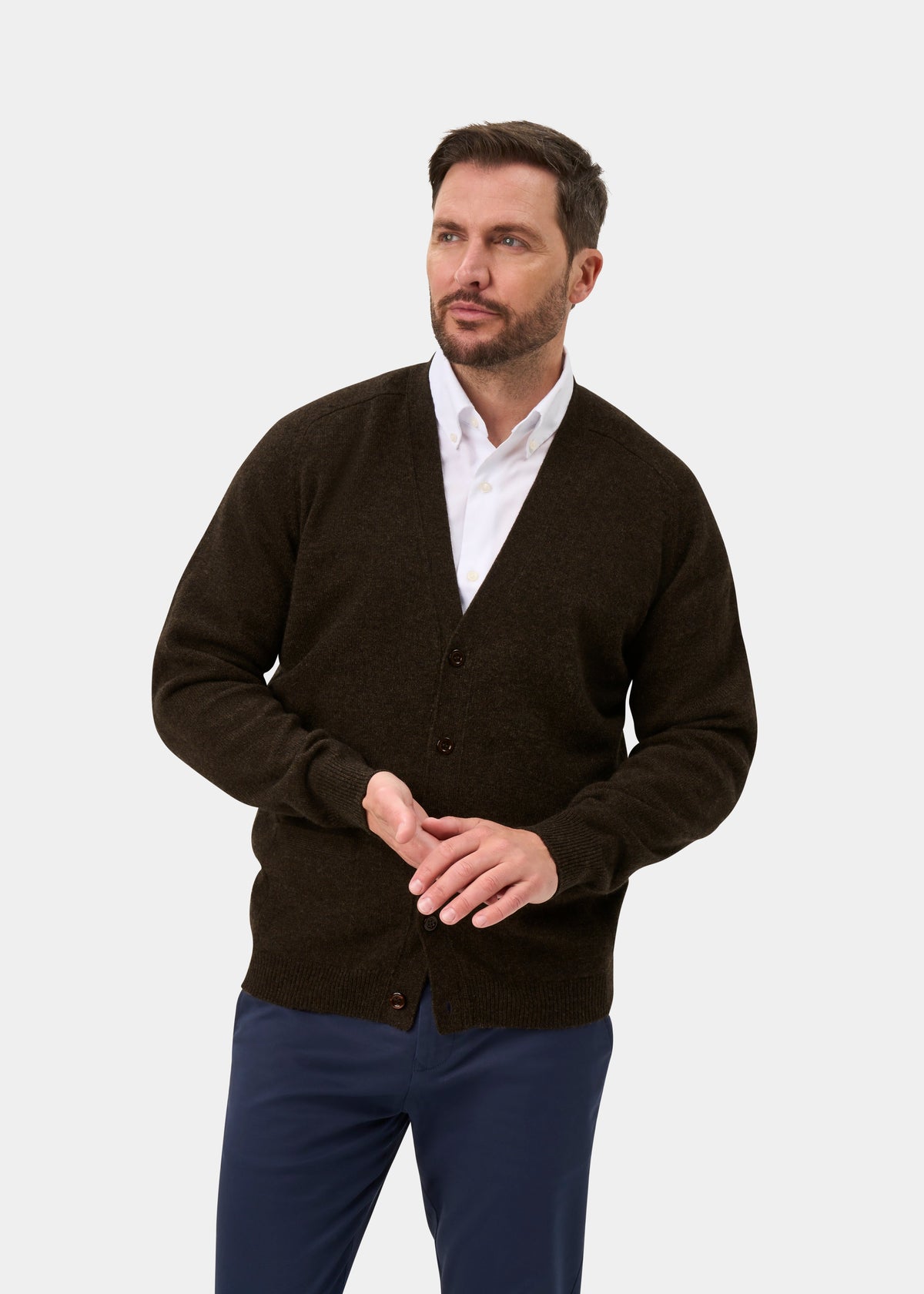 Man wears Cornwall Lambswool V-Neck Cardigan in Cocoa, white shirt, blue pants; stands by a plain light background.
