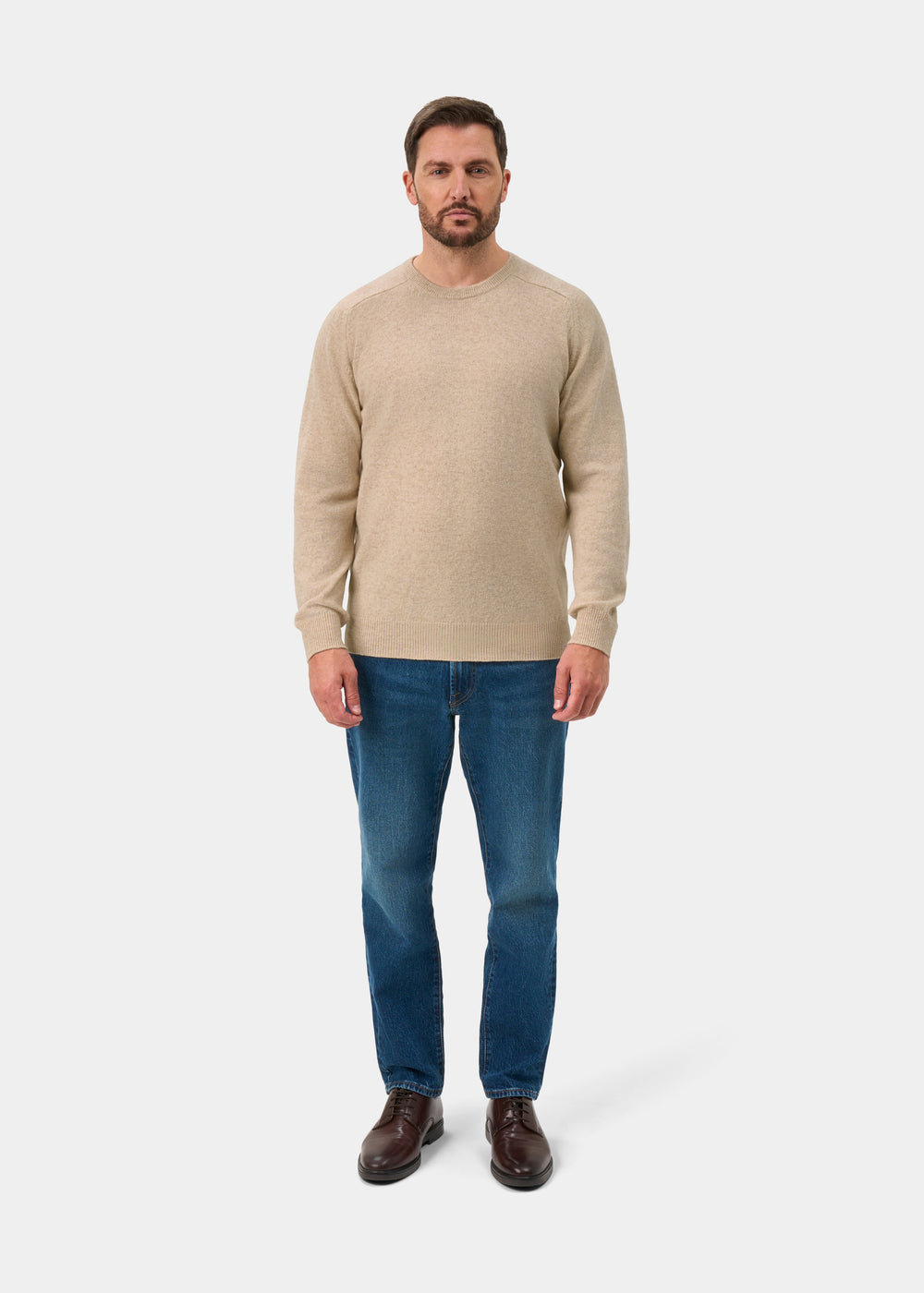 Man in Dorset Lambswool Crew Neck Jumper in Linen, blue jeans, and brown shoes against a white background.