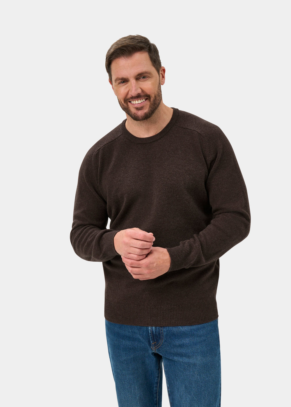 Smiling man wears Dorset Lambswool Crew Neck Jumper in Mocha, classic fit, and blue jeans on white background.