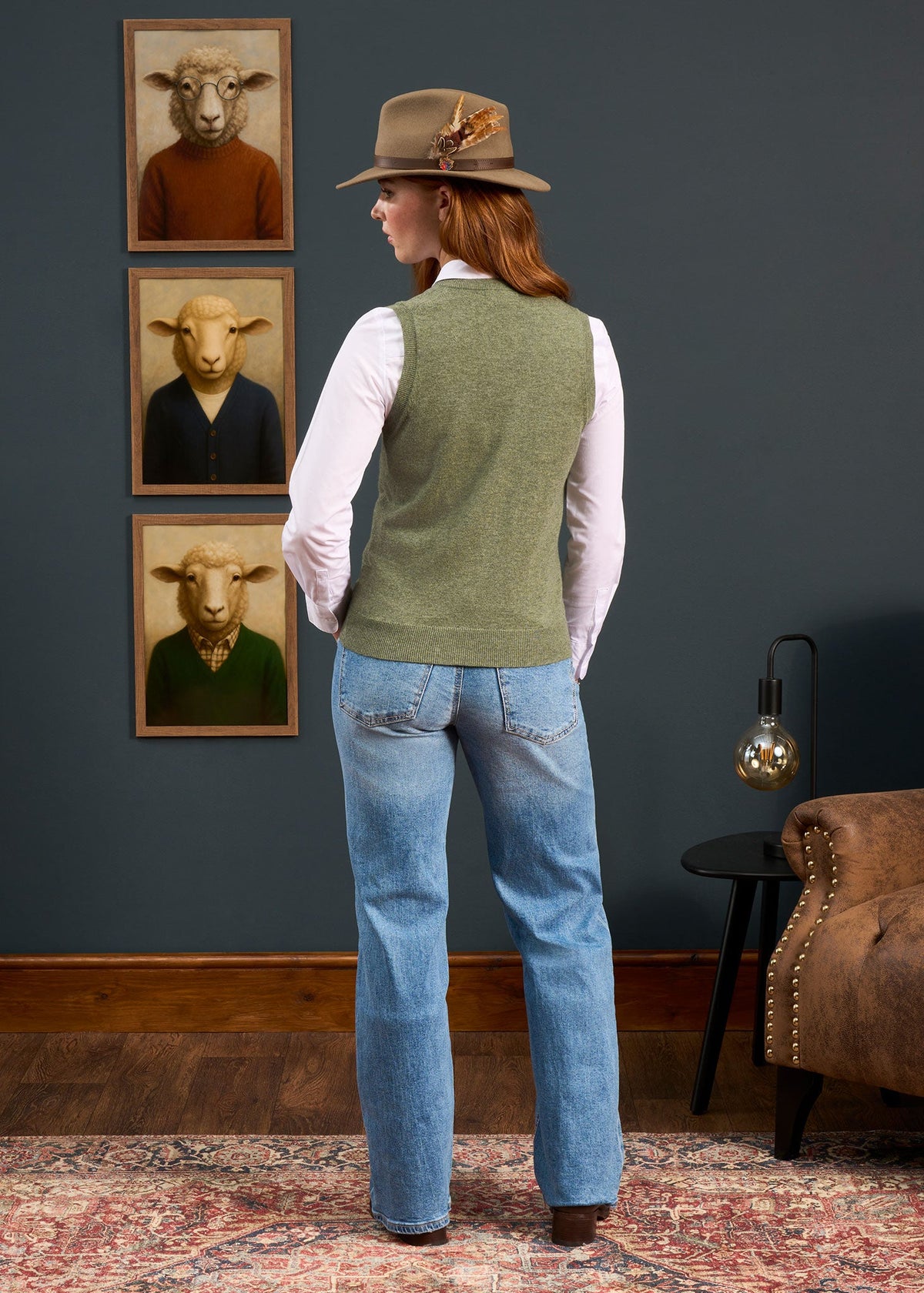 Woman in Erin Wool & Cashmere V-Neck Slipover faces four sheep portraits on a dark wall in a cozy, stylish room.